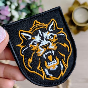 May include: Black embroidered patch featuring a roaring lion's head with a crown. The lion's face is white with orange and yellow accents, set against a black background. The patch is a shield shape, suitable for jackets, bags, or other fabric items.
