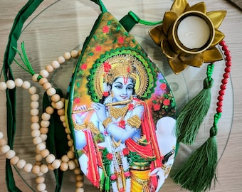 Puede incluir: Una bolsa en forma de hoja con una imagen vibrante de una deidad tocando la flauta. La bolsa tiene una correa y ribete verdes, con un hilo de cuentas y un candelabro de flor de loto dorado. También hay una cadena de cuentas de madera.