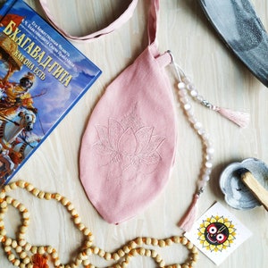 May include: A pink shoulder bag with a lotus flower design, a book with Russian text, and a beaded necklace. The bag has a long strap and a tassel. A small card with a sun design is also visible.