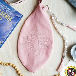 May include: A pink fabric bag with a lotus flower embroidery, a strap, and a drawstring closure. A strand of white beads with a tassel and a wooden bead necklace are also visible. A book with Russian text is in the background.