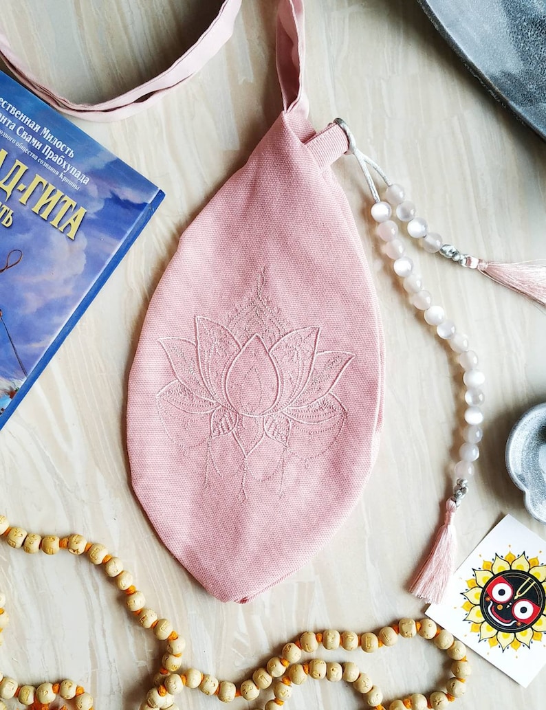 May include: A pink fabric bag with a lotus flower embroidery, a strap, and a drawstring closure. A strand of white beads with a tassel and a wooden bead necklace are also visible. A book with Russian text is in the background.