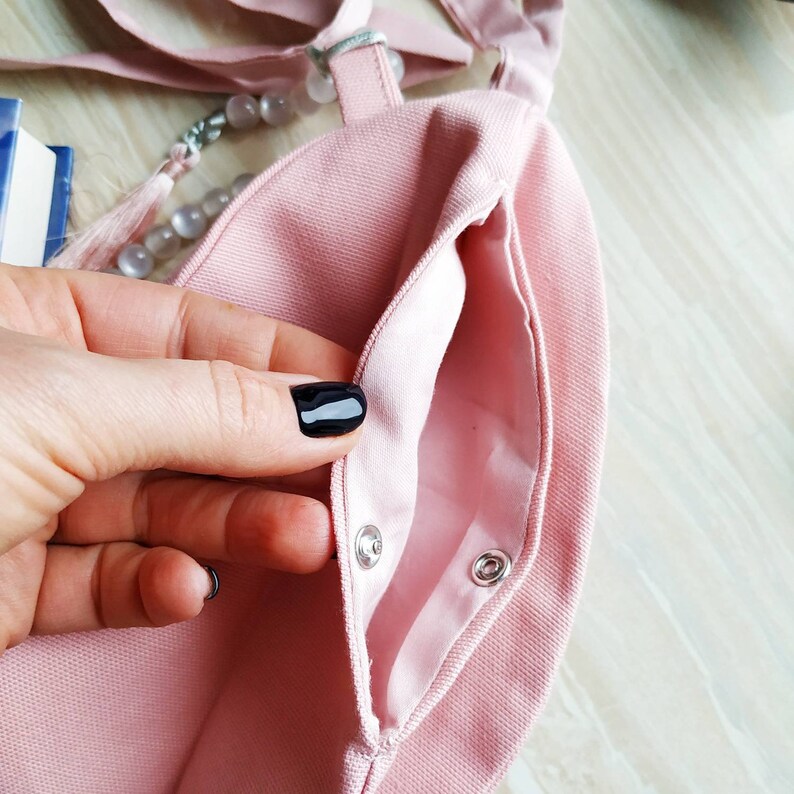 May include: A light pink canvas tote bag with a snap closure. The bag has a matching pink interior and a long strap. A hand with black nail polish is holding the bag open, revealing the inside pocket.