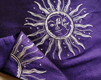 Könnte beinhalten: Lilafarbener Stoff mit einem silbernen gestickten Sonnen- und Mondmotiv. Die Sonne hat ein Gesicht und der Mond hat zwei Gesichter.