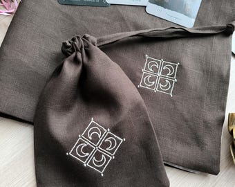 Könnte beinhalten: Zwei braune Leinensäckchen mit weißen gestickten Halbmond-Designs. Die Säcke liegen auf einer weißen Oberfläche.