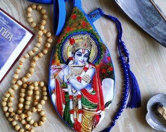 Puede incluir: Una bolsa de tela azul y dorada con una representación de Krishna tocando la flauta. La bolsa tiene un borla azul y está decorada con motivos florales.