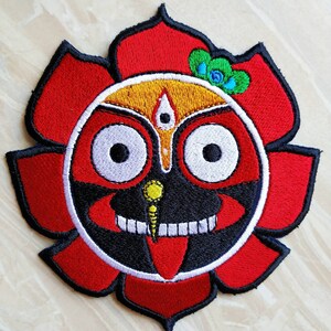 May include: Embroidered patch featuring a red, black, and white deity with large eyes, a red tongue, and a yellow nose. The deity is surrounded by a red lotus flower with green and blue accents.