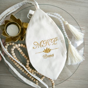May include: A white canvas bag with a gold embroidered lotus flower and the text "M.K.P. Prana". The bag has a drawstring closure and a white tassel.