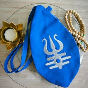 May include: A blue fabric bag with a white embroidered symbol. The symbol is a trident with three prongs and a horizontal line below it. The bag has a long strap and is lying on a glass surface.