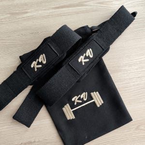 May include: Black weightlifting straps with embroidered "KV" logo and a matching black drawstring bag featuring the same logo and a barbell design. The straps are made of a durable, woven material.
