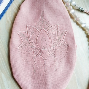 May include: A pink, oval-shaped yoga mat cover with an embroidered lotus flower design. The lotus is detailed with intricate patterns in a lighter shade of pink. The cover is made of a textured fabric.
