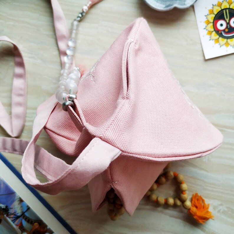 May include: A light pink fabric bag with a triangular shape, featuring a small side pocket and a handle. The bag is adorned with a beaded strap and delicate embroidery. A wooden bead necklace and a small illustration are also visible.