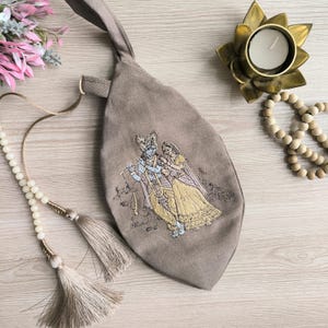 May include: A taupe-colored, leaf-shaped bag with an embroidered image of two figures in traditional attire. The bag has a handle and a gold ring. A beaded necklace with tassels and a brass lotus candle holder are also visible.