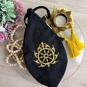 May include: Black fabric bag with a gold embroidered Dharmachakra symbol. The bag has a long strap and is decorated with yellow tassels.
