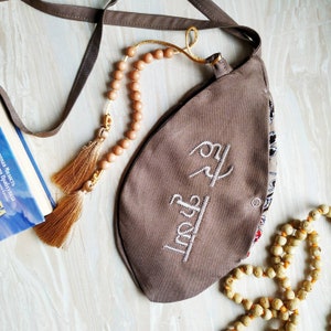 May include: A brown fabric bag with a drawstring closure and a beaded strap. The bag has a white embroidered design with the word "Truth" in Hindi. The bag is decorated with a beaded tassel.