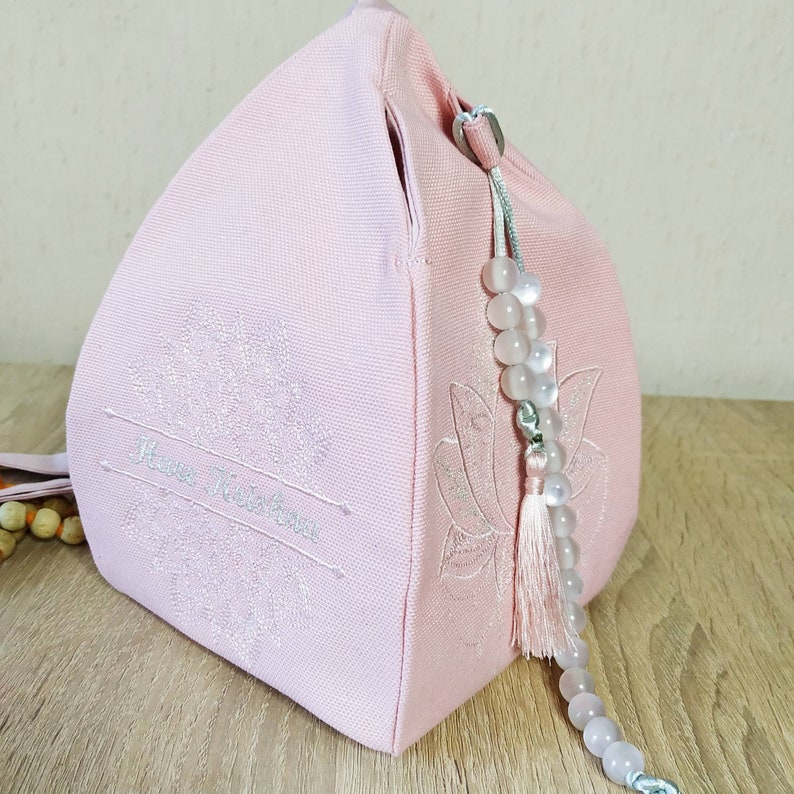 May include: A light pink fabric bag with a lotus flower design embroidered on the side. The bag has a drawstring closure and a beaded tassel. The text "Have Wisdom" is embroidered on the front. The bag is sitting on a wooden surface.