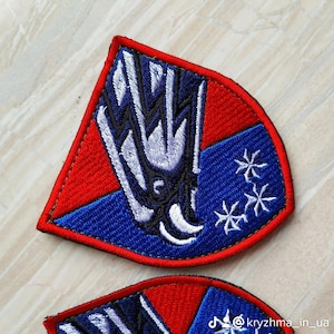 May include: Embroidered patch with a red border and a blue and white design. The patch features a stylized emblem with a central figure and three white star-like shapes. The patch is likely for clothing or accessories.