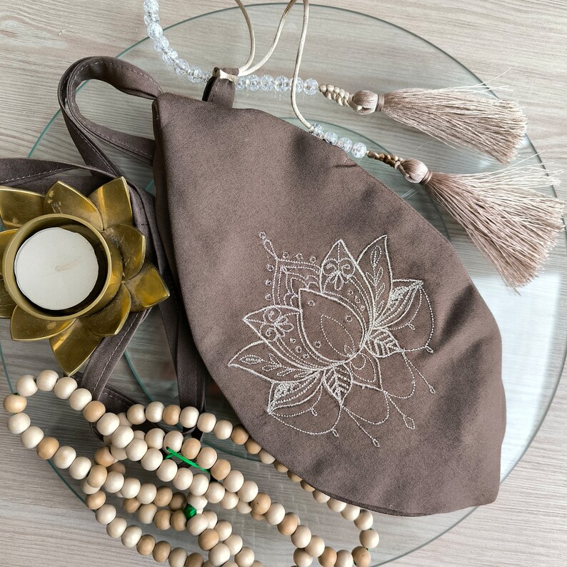 May include: A brown fabric pouch with a white embroidered lotus flower design. The pouch has a drawstring closure and is surrounded by wooden beads and a white tassel.