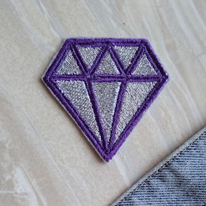Könnte beinhalten: Ein aufbügelbarer Diamant-Patch in Lila und Silber. Der Diamant ist mit Lila umrandet und mit silbernem Glitzer gefüllt.