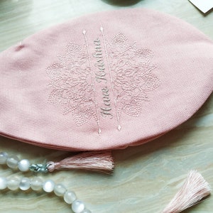 May include: A light pink prayer cap with embroidered floral design and the words "Hare Krishna". A strand of white beads with a tassel is in the foreground. The cap is made of a soft fabric.
