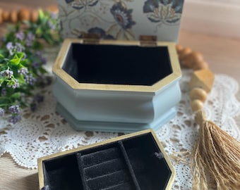 Puede incluir: Una caja de joyería de madera azul claro con un patrón floral en la tapa. La caja tiene un interior de terciopelo negro y un soporte para anillos con múltiples ranuras.