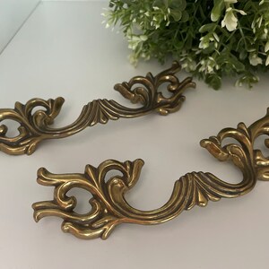 May include: Two ornate brass drawer pulls with a decorative, scroll-like design. The pulls are in a classic, vintage style.