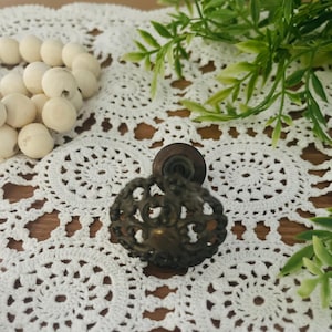 May include: A vintage-style, ornate, brass cabinet knob with a detailed floral design. The knob is on a white crocheted doily.