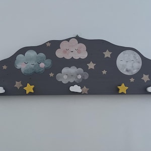 Puede incluir: Un colgador de pared de madera gris oscuro con forma de nube con una luna y estrellas. El colgador tiene cinco ganchos, cada uno con forma de nube. Las nubes están pintadas en varios tonos de azul, rosa y gris, y tienen caras sonrientes. La luna es blanca con una cara sonriente. Las estrellas son doradas.