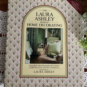 Può includere: Il libro "Laura Ashley Book of Home Decorating", un libro verde e bianco con un motivo floreale sulla copertina. Il libro presenta una foto di una stanza con una poltrona bianca e un tavolo di legno. Il testo sulla copertina recita "The Laura Ashley Book of Home Decorating" e "Nuova edizione riveduta e corretta".