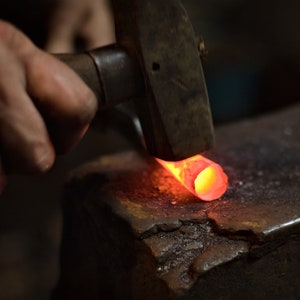 May include: A blacksmith forges metal with a hammer on an anvil. The metal is glowing red-orange from the heat. The scene is dimly lit, highlighting the intense process of metalworking.