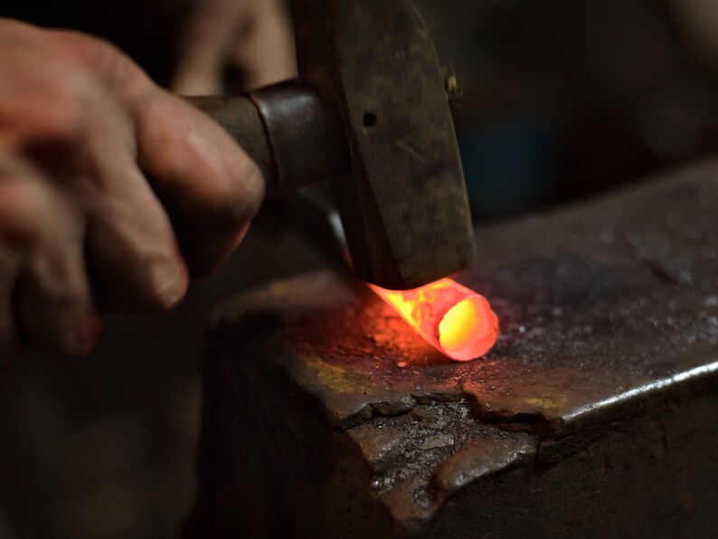 May include: A blacksmith forges metal with a hammer on an anvil. The metal is glowing red-orange from the heat. The scene is dimly lit, highlighting the intense process of metalworking.