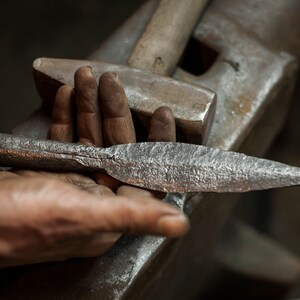 May include: A hand holds a silver metal spearhead with a rough, hammered texture. The spearhead is resting on a metal anvil.