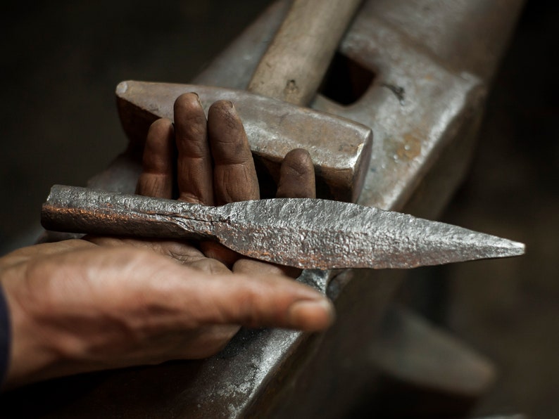 May include: A hand holds a silver metal spearhead with a rough, hammered texture. The spearhead is resting on a metal anvil.
