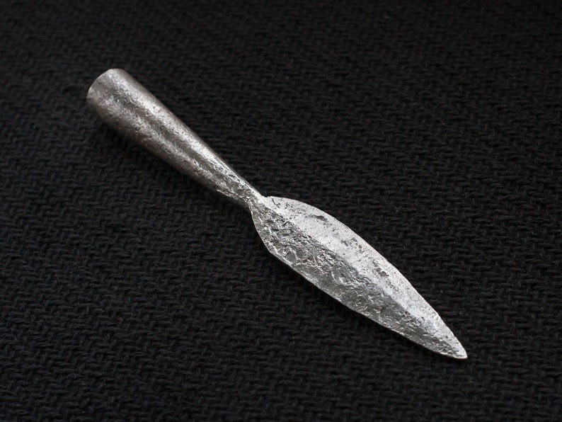 May include: A silver-colored spearhead with a textured surface. The spearhead has a pointed blade and a cylindrical base. The spearhead is likely made of metal and is set against a black background.