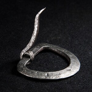 May include: A silver metal belt buckle with a curved, pointed tongue. The buckle is made from a single piece of metal and has a simple, rustic design.