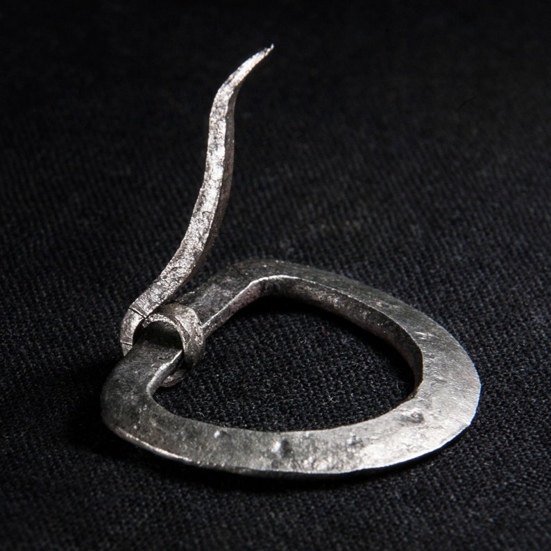 May include: A silver metal belt buckle with a curved, pointed tongue. The buckle is made from a single piece of metal and has a simple, rustic design.