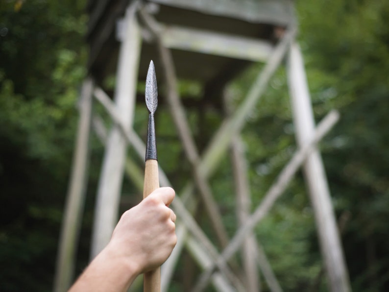 May include: A spear with a metal arrowhead and a wooden shaft is held up against a blurred background of a wooden structure and green foliage. The spearhead is silver, and the shaft is light brown.