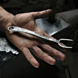 Könnte beinhalten: Handgeschmiedete Metallgabel mit strukturierter Oberfläche, mit einer Schlaufe am Ende des Griffs und zwei Zinken. Die Gabel wird in einer Hand gehalten, was die Handwerkskunst des Schmiedens zeigt. Der Hintergrund umfasst einen Amboss und andere Werkzeuge.
