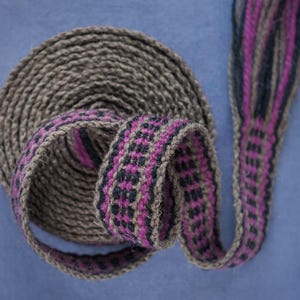 Op de afbeelding: Een geweven band met een bruine basis en een geometrisch patroon in roze en zwart. De band is opgerold en gevouwen.