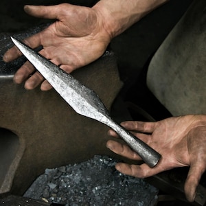 May include: A hand-forged, spearhead with a sharp, pointed tip. The metal has a textured, hammered appearance, and the spearhead is held against a dark background, suggesting a blacksmith's workshop. The spearhead is a historical or decorative item.