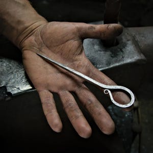 May include: A hand-forged metal tool with a pointed end and a looped handle. The tool is silver in colour and appears to be made of iron or steel. The background shows a blacksmith's workspace, suggesting a handmade craft.
