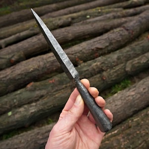 Puede incluir: Una punta de lanza de acero forjado a mano con una punta afilada y puntiaguda. El metal tiene un acabado gris oscuro texturizado. La punta de lanza se sostiene contra un fondo de troncos apilados.