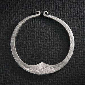 Peut inclure: Un collier en métal argenté martelé, de forme circulaire. Le collier présente une surface texturée et un petit détail pointu en bas. Les extrémités du collier sont incurvées vers le haut, formant de petites boucles.