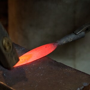 May include: A close-up of a red-hot metal rod being hammered on an anvil. The metal is glowing orange and is being shaped by the hammer.