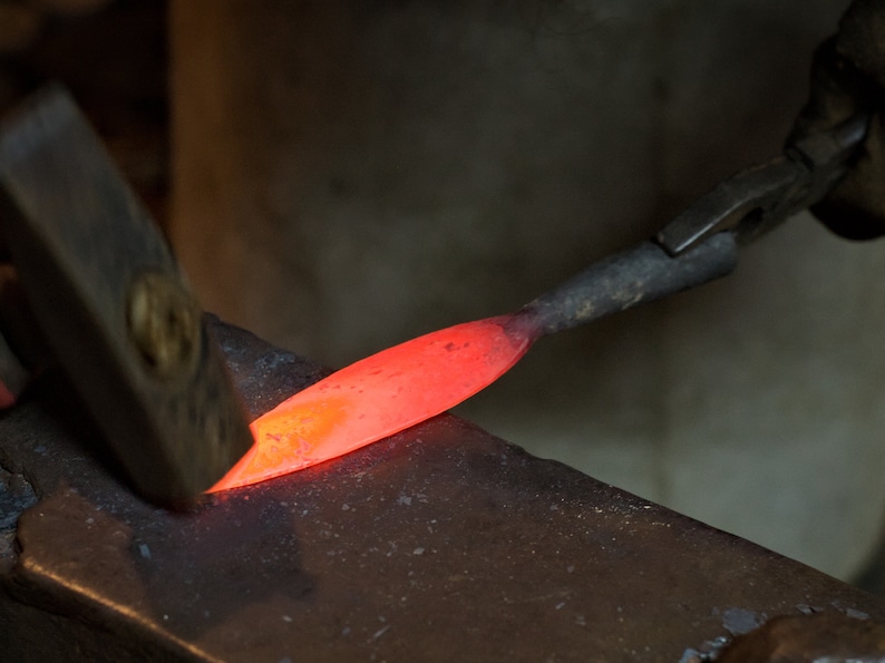 May include: A close-up of a red-hot metal rod being hammered on an anvil. The metal is glowing orange and is being shaped by the hammer.