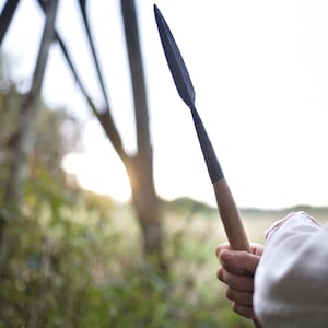 Może przedstawiać: Zbliżenie na osobę trzymającą wł&oacute;cznię z drewnianym trzonkiem i ciemną metalową końc&oacute;wką. Wł&oacute;cznia jest skierowana w stronę nieba, a tło jest rozmyte.