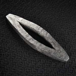 Puede incluir: Un objeto plateado y alargado con una superficie texturizada y una abertura central. El objeto tiene una forma ovalada simétrica con extremos puntiagudos. El fondo es una tela oscura y texturizada.
