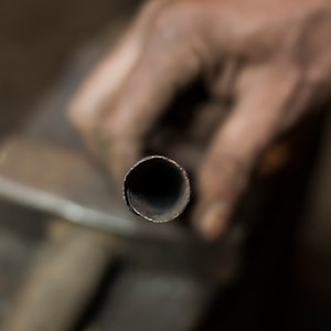 May include: Close-up of a metal pipe being held by a hand. The pipe is round and has a dark gray finish.