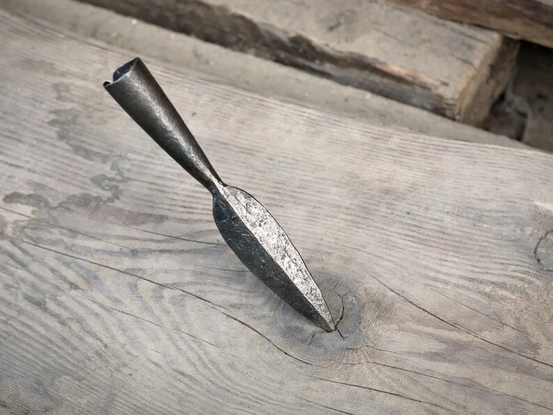May include: A hand-forged, antique spearhead with a tapered shaft and a leaf-shaped blade. The metal has a dark, aged appearance, resting on a weathered wooden surface. The spearhead is a historical artifact.