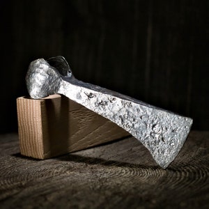 Könnte beinhalten: Eine silberne Axt mit einem abgerundeten Kopf und einer strukturierten Oberfläche. Die Axt liegt auf einem Holzblock.