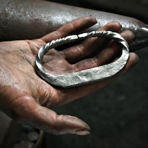 May include: A silver bracelet with a twisted rope design and a hammered texture. The bracelet is held in a hand, highlighting the craftsmanship. The metal has a matte finish. The bracelet's design is visible.